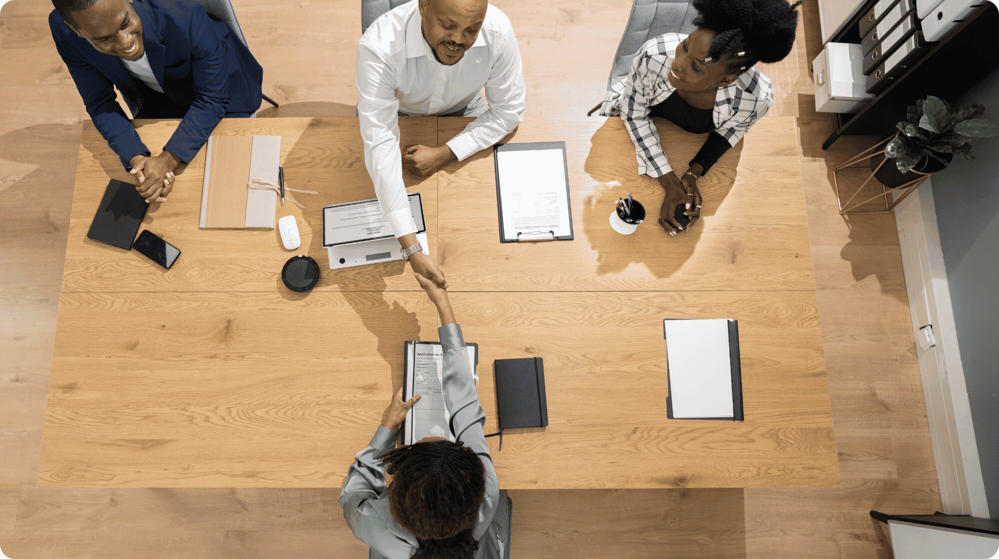 Overhead view of table with three interviews sitting opposite the candidate. The interviewer sat in the middle is shaking the candidate's hand.