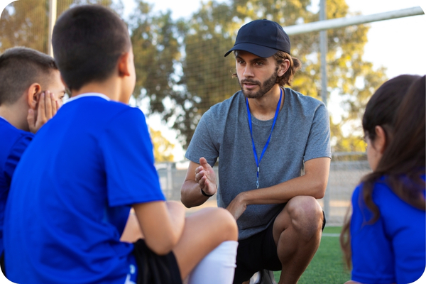 A coach in a basketball cap is crouching down briefing a children's sports team