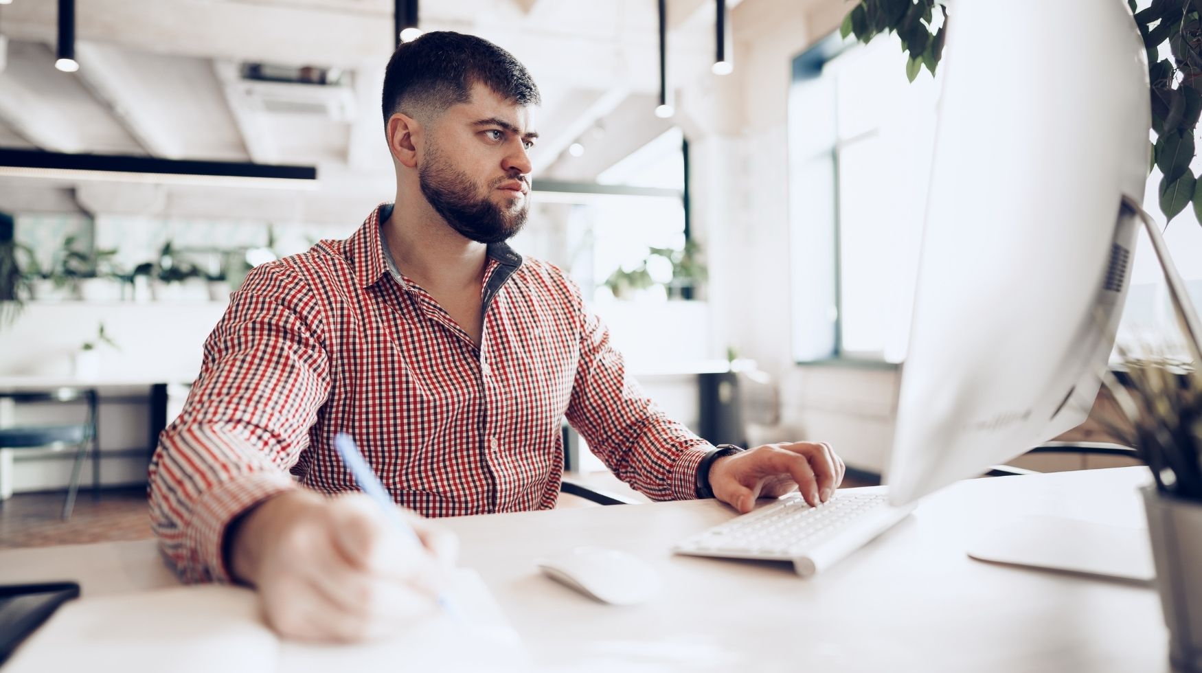 A man is sat at a desk looking at a computer and making notes in a paper pad.